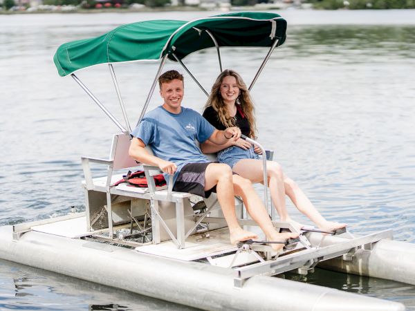 A smiling couple relaxes on a pedal boat with a green canopy, enjoying a sunny day on the water.