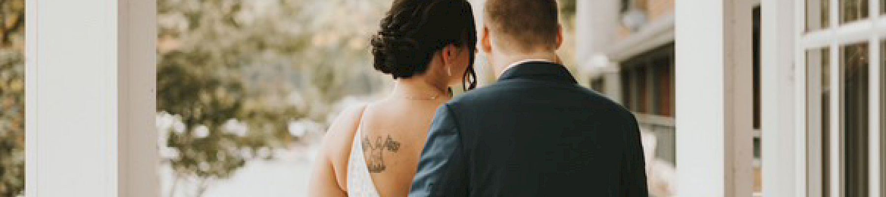 A couple stands together on a balcony, woman in a white dress with tattoo, man in a dark suit, overlooking a scenic view.