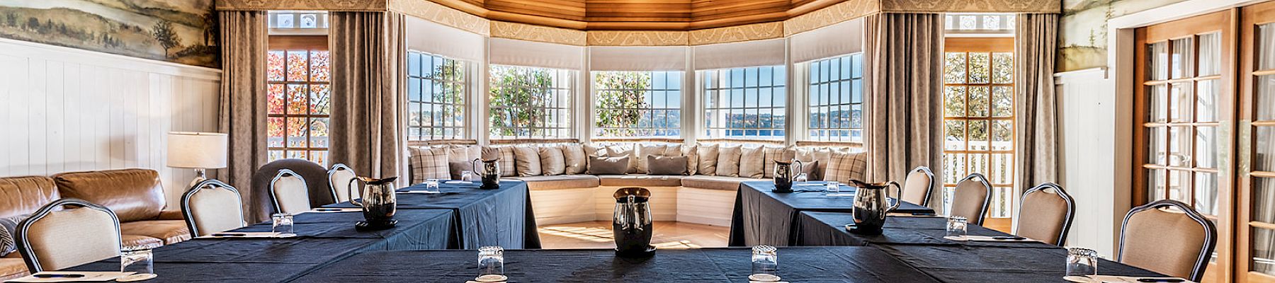 A conference room with U-shaped seating, tables with black cloths, and a wooden ceiling. Large windows offer natural light and scenic views.