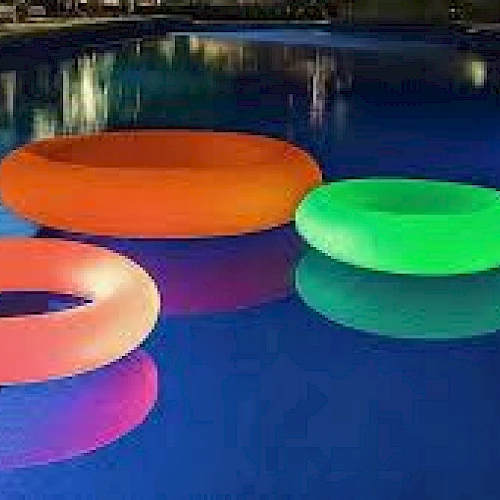 The image shows a pool with colorful, glowing inflatable rings floating on the water, creating a vibrant nighttime scene.
