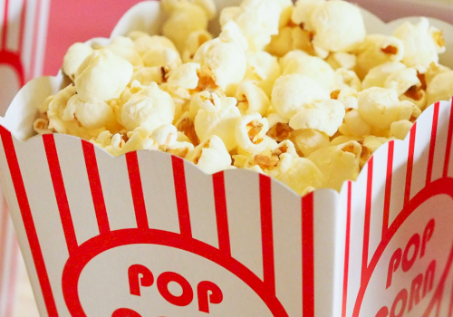 A tub of popcorn in a red-and-white striped popcorn box labeled &ldquo;POP CORN,&rdquo; with other snacks blurred in the background.