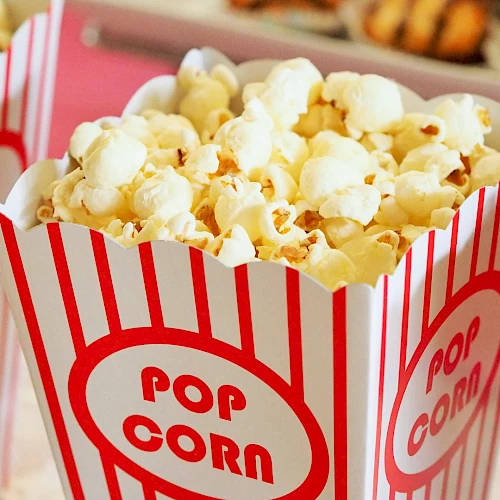 A tub of popcorn in a red-and-white striped popcorn box labeled &ldquo;POP CORN,&rdquo; with other snacks blurred in the background.