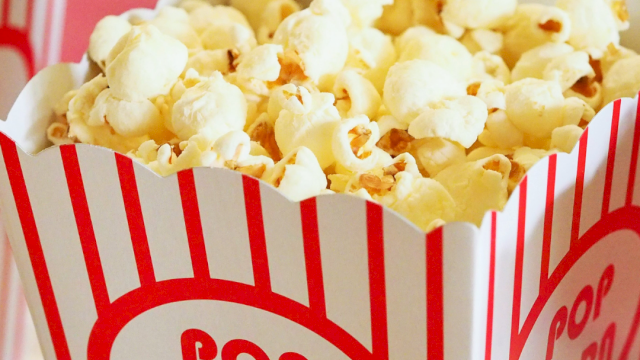 A tub of popcorn in a red-and-white striped popcorn box labeled “POP CORN,” with other snacks blurred in the background.