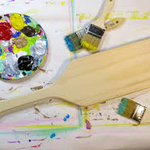 A paint palette with colorful paints, a wooden board, and three paintbrushes on a paint-splattered surface end the scene.