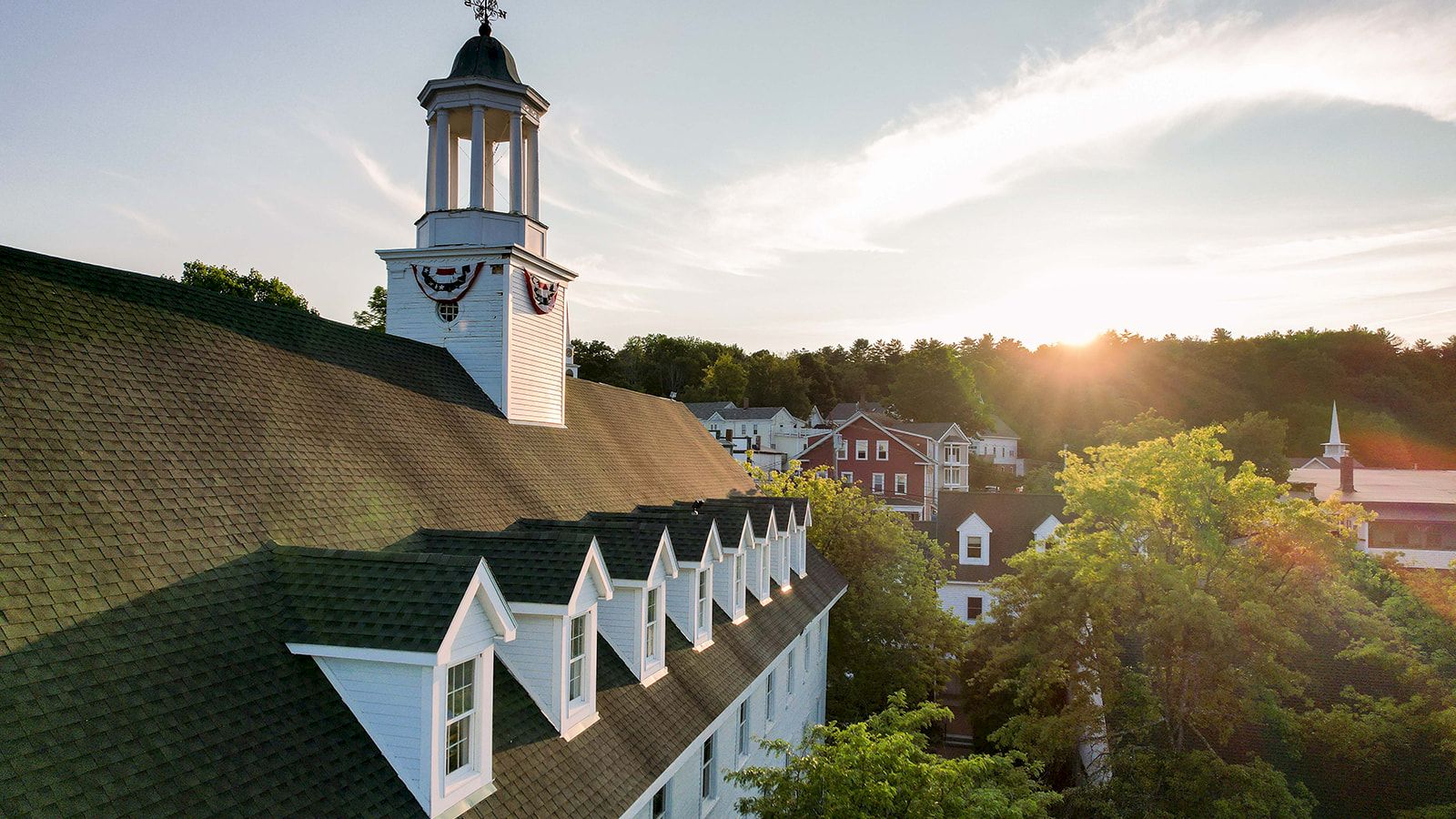 A sunlit resort hotel roof with dormer windows, a white cupola, and a town skyline beyond, basking in warm evening light.