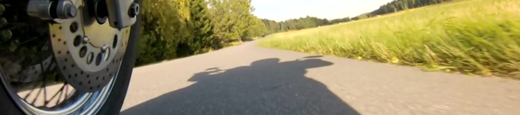 A motorcycle wheel on a country road, casting a shadow. The road is surrounded by green fields and trees under a clear blue sky.