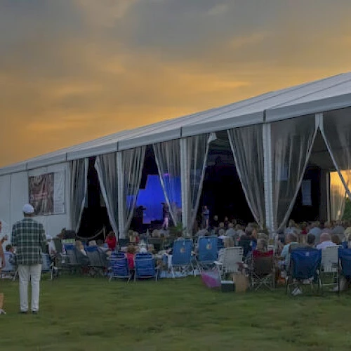 A crowd seated outdoors in front of a white tent enjoys an event during a colorful sunset.