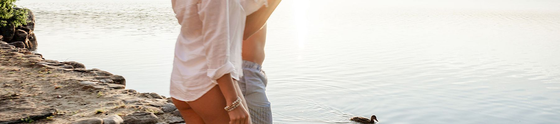 Two people are walking barefoot by a lakeside at sunset with a duck swimming nearby.