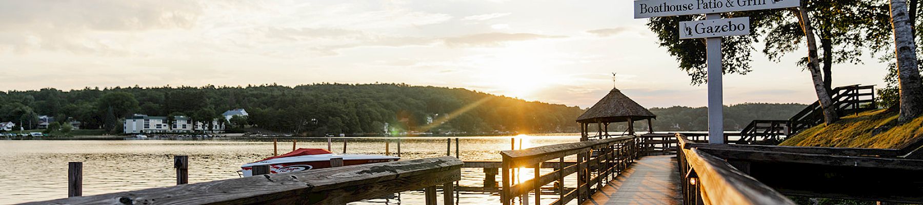 A wooden boardwalk leads to a gazebo by the water at sunset, with signposts pointing to various destinations.