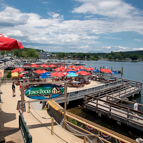 A waterfront dining area with colorful umbrellas, a sign reading "Town Docks," and a picturesque view of the lake and sky.