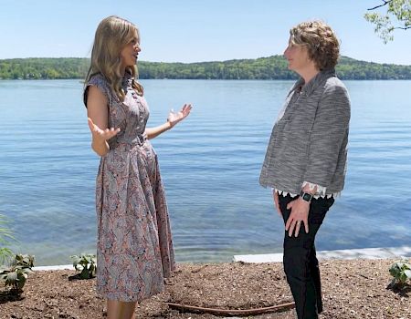 Two people are talking by a lakeside, surrounded by greenery and a clear blue sky.