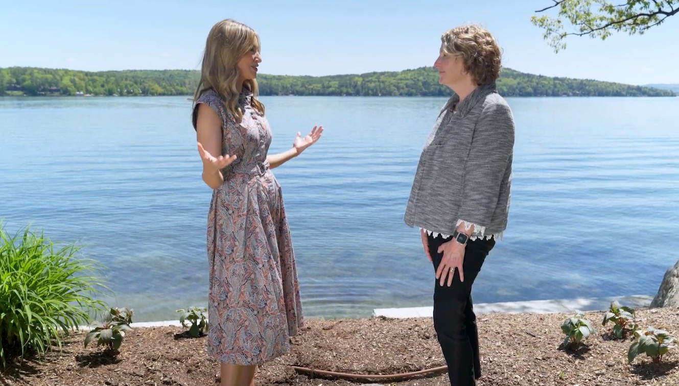 Two people are talking by a lakeside, surrounded by greenery and a clear blue sky.