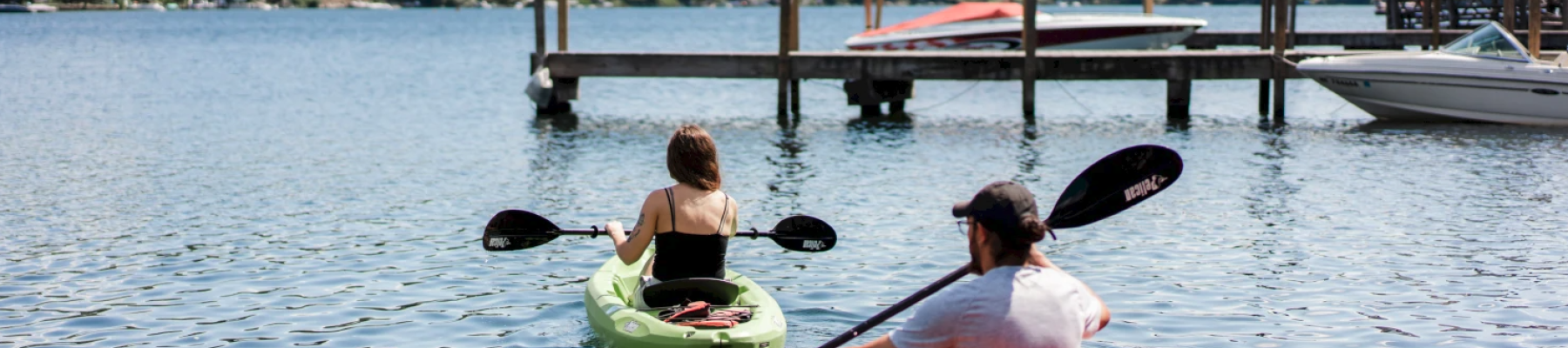 Two people are kayaking on a lake near a wooden dock, with trees and clouds in the background, creating a serene outdoor scene.