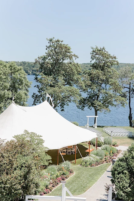 A white tent in a scenic lakeside garden setting, surrounded by trees and flowers, with a walkway leading to the water.