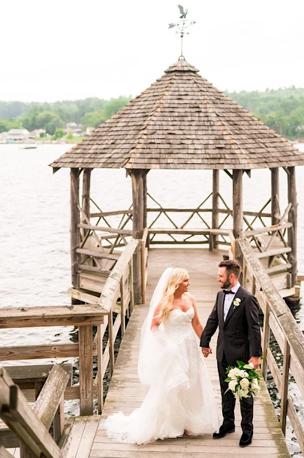 A couple in wedding attire stands on a wooden dock by the water, under a gazebo, holding hands.