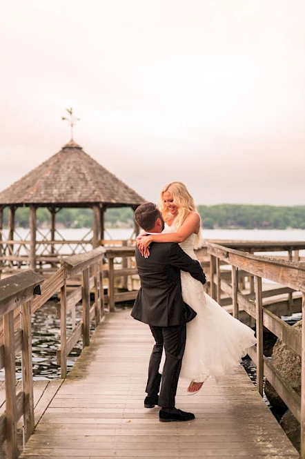 A couple embraces on a wooden dock by the water, with a gazebo in the background under a cloudy sky.