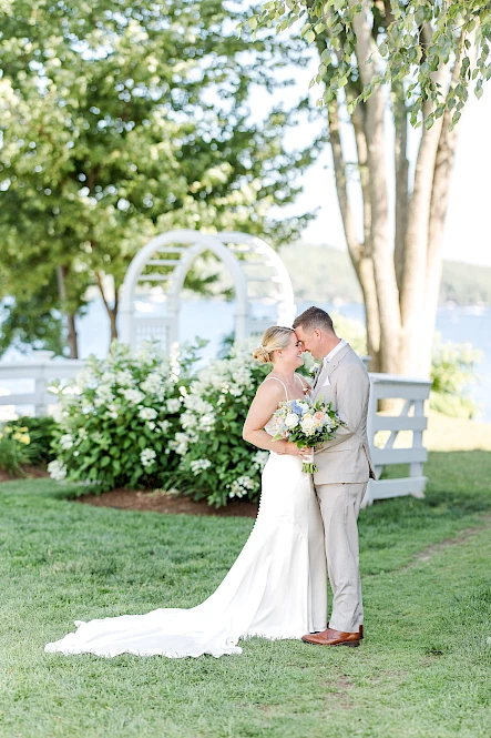 A couple is kissing outdoors in a garden setting, dressed in wedding attire, with greenery and flowers around them on a sunny day.