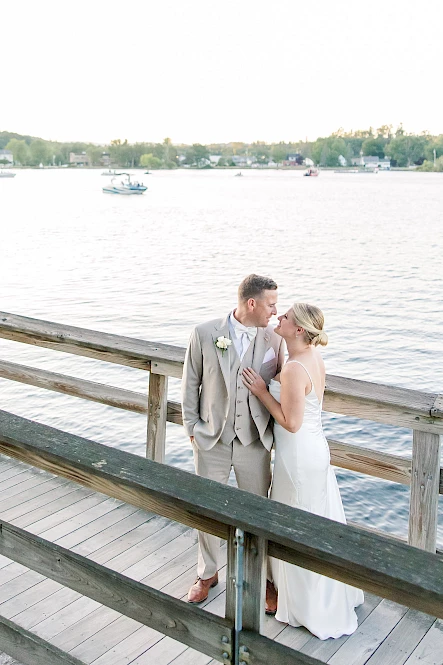 A couple is embracing on a wooden pier by a lake, with boats and trees in the background, dressed in formal wedding attire.