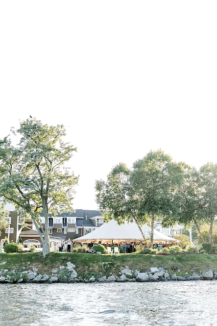 A waterfront scene with a white tent, trees, and a building in the background, viewed from across a body of water.