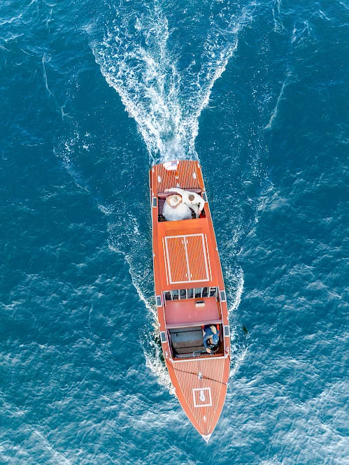 Aerial view of an orange boat cutting through blue sea, with passengers on board and white wake behind it.