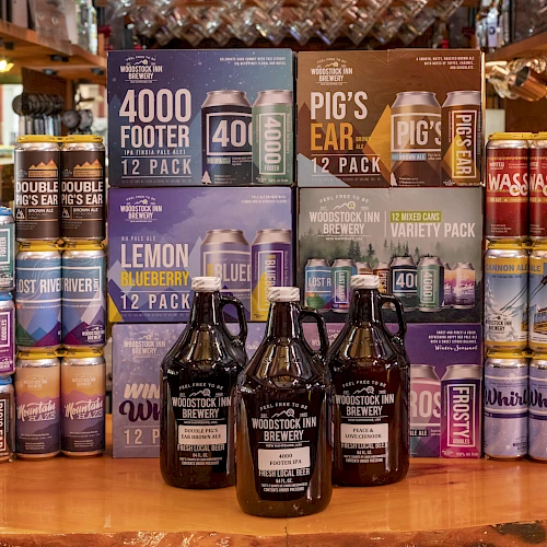 A display of assorted craft beer and seltzer packs behind bottles, with colorful cans stacked around and a few dark beer bottles in front, all on a wooden surface.