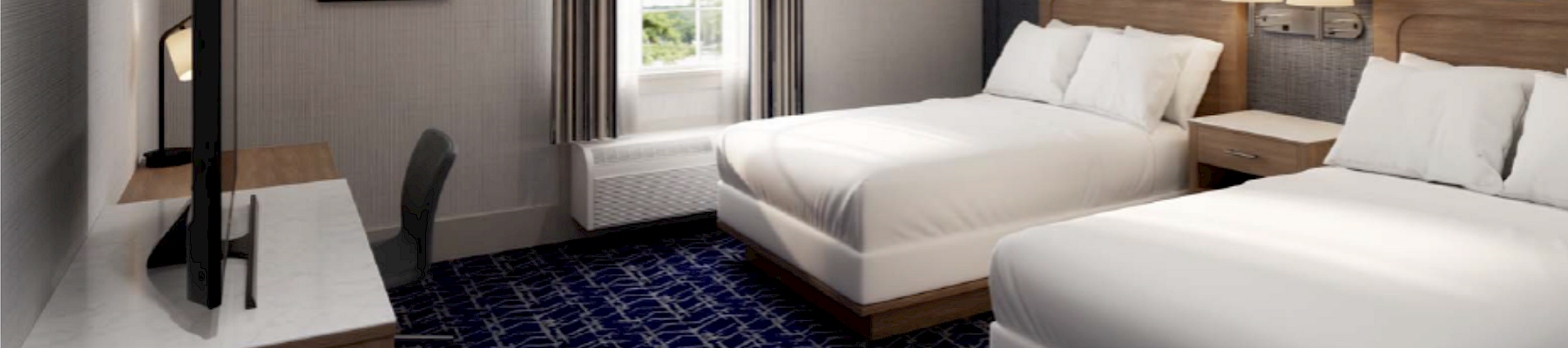 A hotel room with two double beds, a desk and chair, a wall TV, blue patterned carpet, a window, and bedside lamps; logo for Mill Falls Resort appears in the corner.