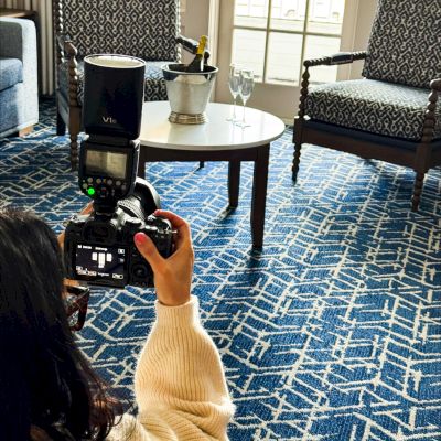 A hotel room scene with a patterned blue carpet, a photographer taking images of furniture and balcony lake view in the background.