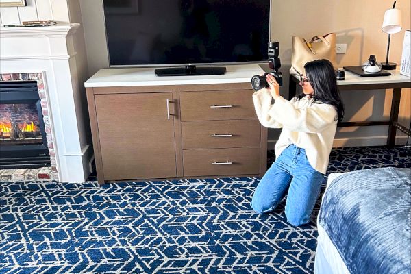A photographer sits on a blue patterned carpet in a hotel room, with a TV stand, couch, and fireplace in the background.