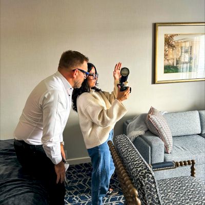 Two people looking through a camera indoors, in a cozy hotel room with a sofa, and sunlight streaming in.