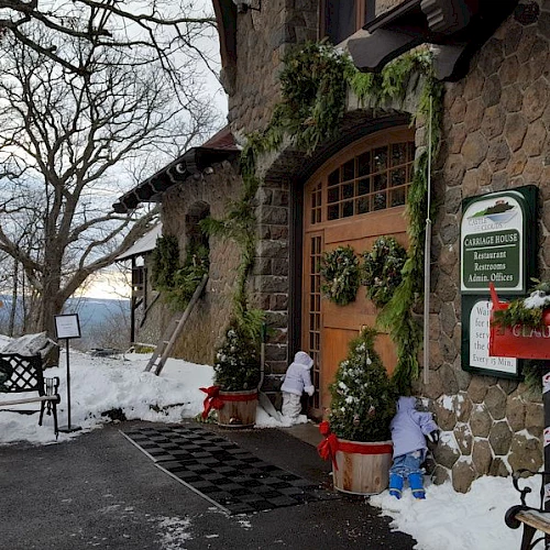 A stone lodge in a snowy scene with wreaths, potted evergreens, benches, and a red mailbox near a wooden door; kids in blue jackets approach.