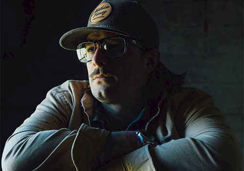 A man wearing a cap and glasses sits with arms crossed, moody lighting and a dark background. He looks contemplative.