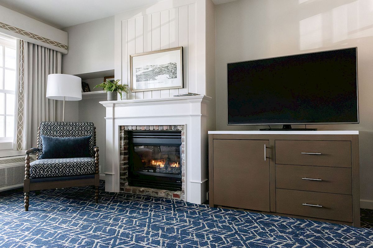 A cozy room features a fireplace, an armchair, a TV on a cabinet, and a patterned blue carpet, creating a comfortable atmosphere.