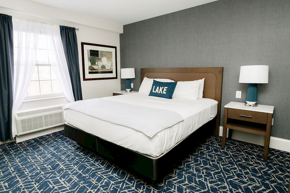 A neatly arranged bedroom with a double bed, two bedside tables, lamps, window with curtains, and a patterned carpet is shown.