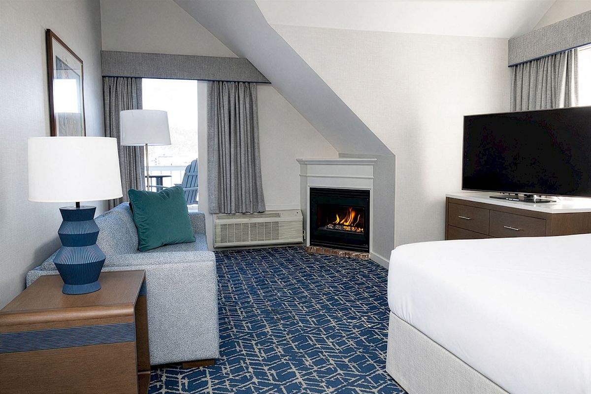 A cozy hotel suite with a bed, sitting area, blue patterned carpet, fireplace, flat-screen TV, and a lamp-lit nightstand in soft neutral tones.