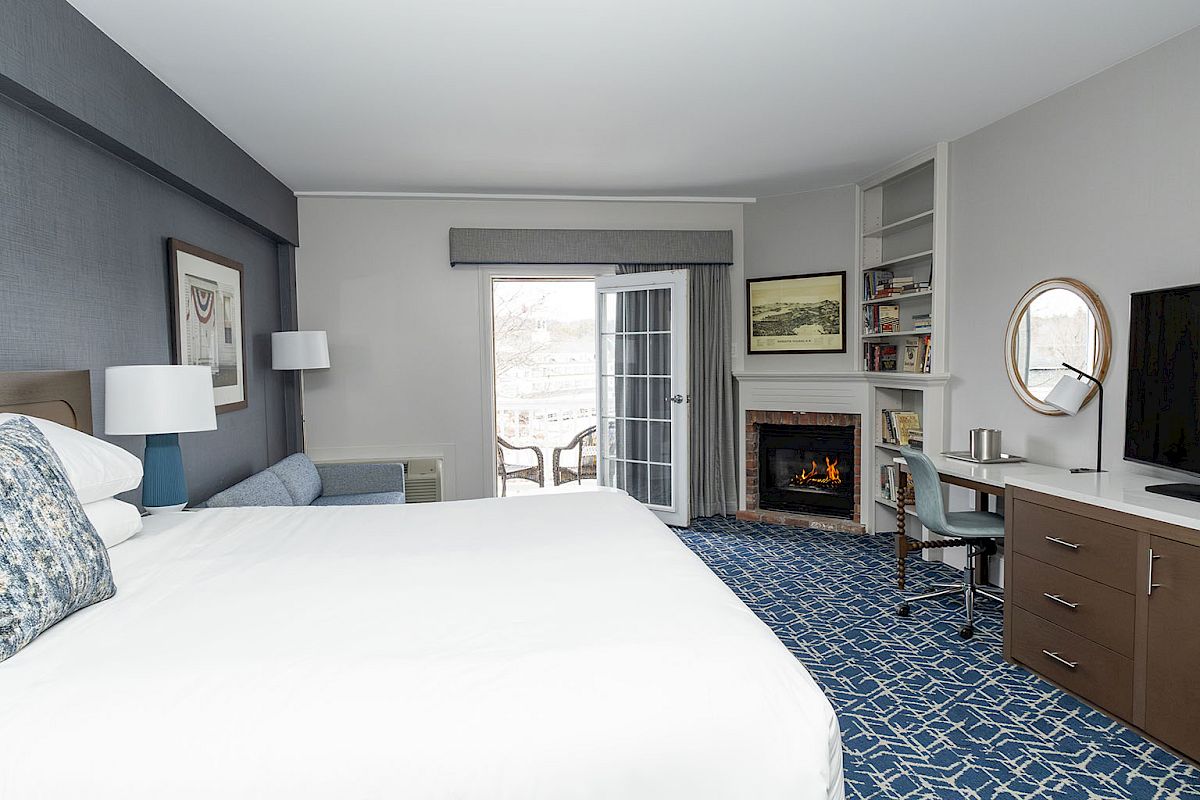 A hotel room with a large bed, blue carpet, desk, fireplace, and sliding door to a balcony, neat and modern with neutral tones.