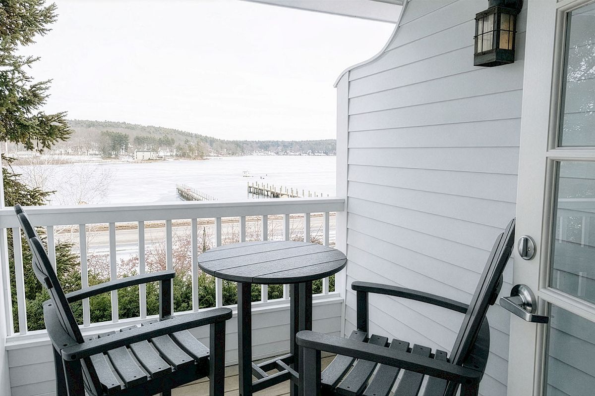 A cozy balcony scene overlooking a frozen lake with a small round table and two black chairs. The porch is white with a lantern by the door.