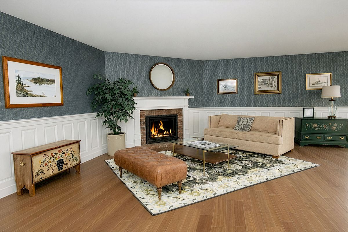 A cozy hotel lobby with a lit fireplace, beige sofa, patterned area rug, tufted leather ottoman, wooden end table, potted plant, and blue wallpapered walls.