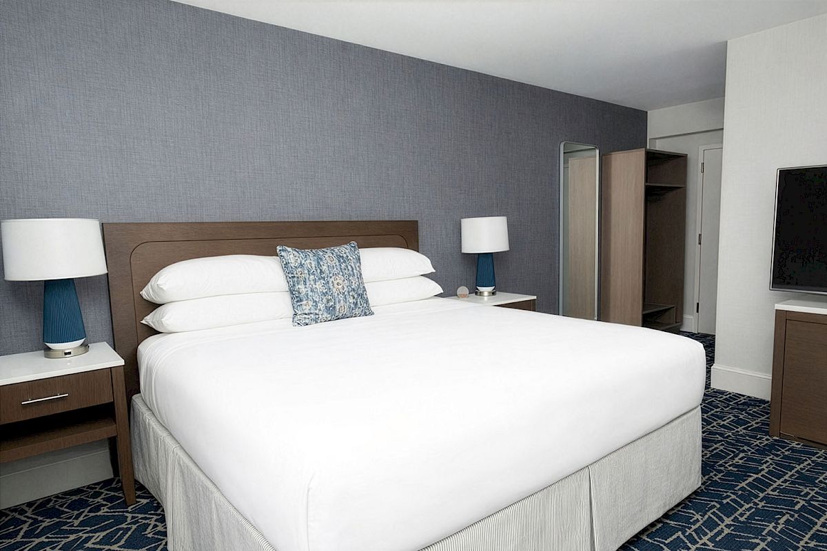 A hotel bedroom with a large white bed, two matching nightstands with lamps, a blue patterned carpet, a wall TV, and a dresser in neutral tones.