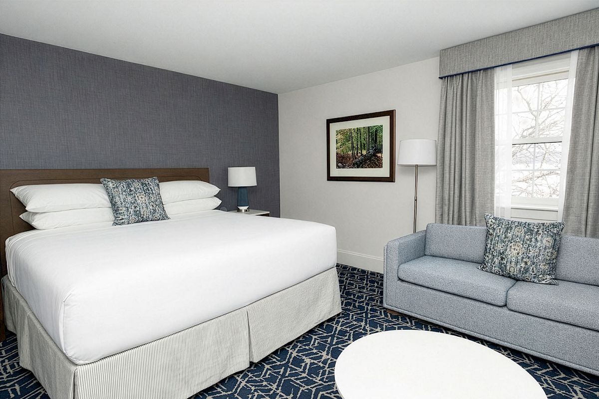 A hotel room featuring a large bed with white linens, a blue accent wall, a sofa, two lamps, a round white table, and a window with curtains.