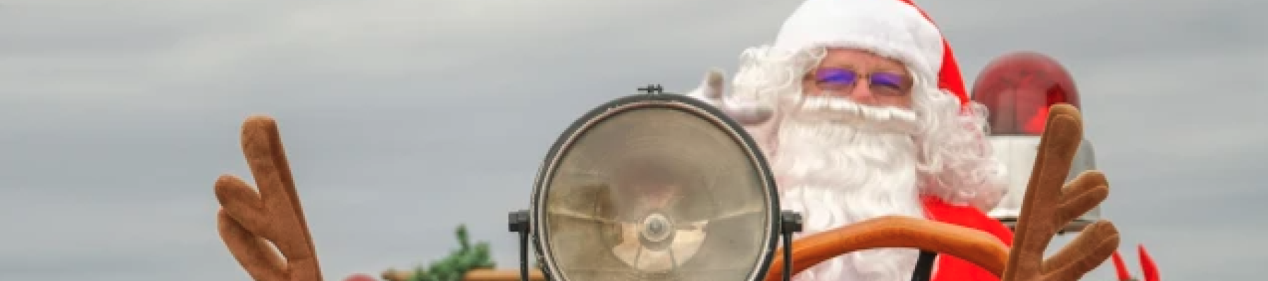 Santa driving a red antique car, wearing a hat, waving with a garland and festive decor on the front—holiday parade vibes.