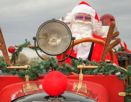 Santa driving a red antique car, wearing a hat, waving with a garland and festive decor on the front—holiday parade vibes.