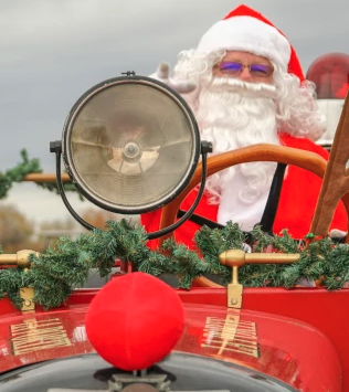 Santa driving a red antique car, wearing a hat, waving with a garland and festive decor on the front—holiday parade vibes.