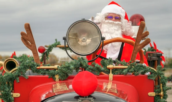 Santa driving a red antique car, wearing a hat, waving with a garland and festive decor on the front—holiday parade vibes.