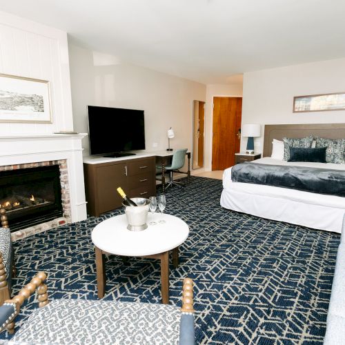 The image shows a hotel room with a bed, a TV, a fireplace, chairs, and a table on a patterned carpet.