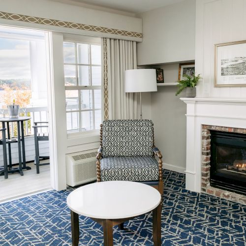 The image shows a cozy living room with a fireplace, chair, and round table, leading to a balcony with a scenic view through a glass door.