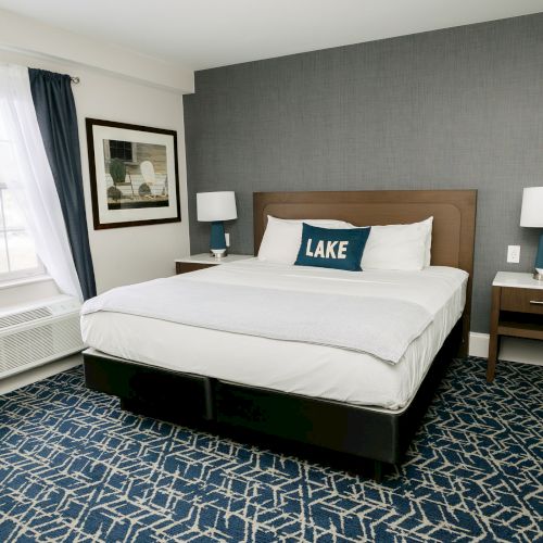 A modern hotel room with a large bed, decorative pillow, nightstands with lamps, patterned carpet, and a window with curtains.