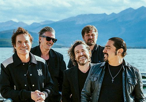 A group of five people standing together outdoors, in front of a scenic backdrop with mountains and water, all smiling.