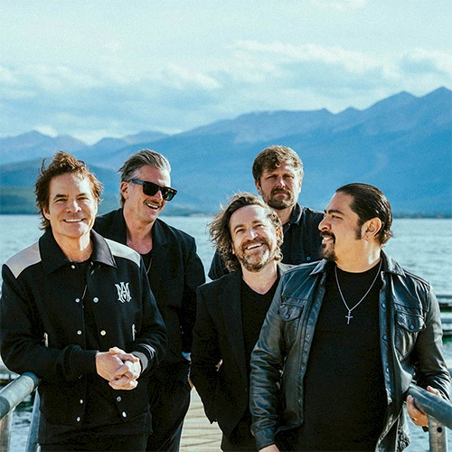 A group of five people standing together outdoors, in front of a scenic backdrop with mountains and water, all smiling.