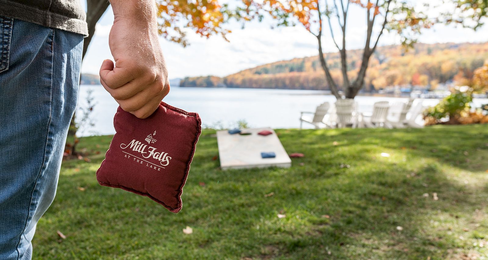 A person holding a red Cornhole bag near a lawn with a cornhole board by a lake and fall trees in the background.