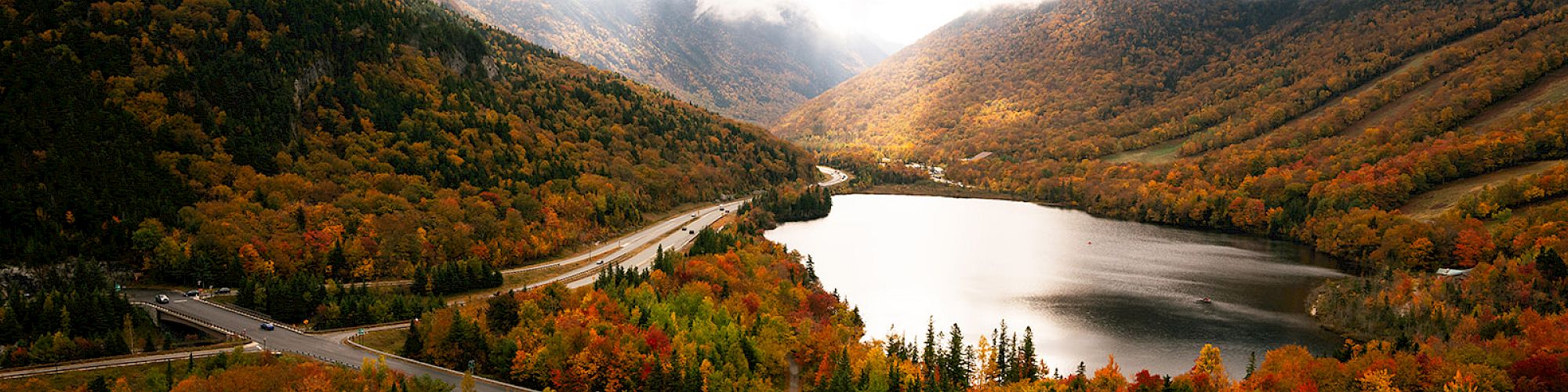 A scenic landscape with a winding road, a lake, and autumn-colored trees nestled between foggy mountains.